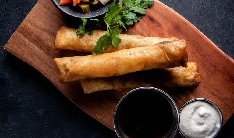 Three crispy tailpipes on a wooden board, garnished with parsley, accompanied by bowls of dipping sauces and sautéed vegetables.